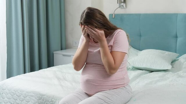 Sad crying pregnant woman suffering from depression sitting on bed and holding her head. Concept of maternal and pregnancy depression.