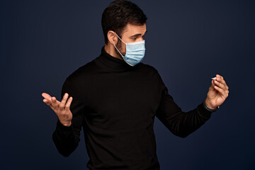 Young man in black sweater, in sterile face mask feeling sick or dizzy, hold thermometer, isolated...