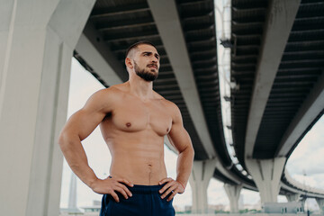 Fototapeta na wymiar Athletic male fitness model keeps hands on waist poses with muscular perfect naked body, looks seriously into distance, does sport exercises outdoor, focused somewhere, goes in for sport regularly