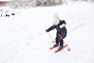 Children's feet in red plastic skis with sticks go through the snow from a slide-a winter sport, family entertainment in the open air. A little girl glides down the slope from an early age. Copy space