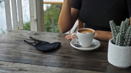 uropean girl sits at a table in a public place with a cup of coffee and nearby to her on a wooden table is a black protective mask