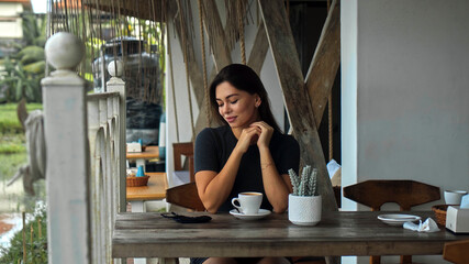 European girl sits at a table in a public place with a cup of coffee on a wooden table and looking on protective mask on a table