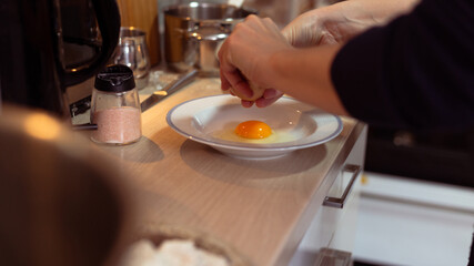 Fototapeta na wymiar Close up shot of Caucasian woman cracking an egg in white plate. Housewife preparing breakfast in kitchen.