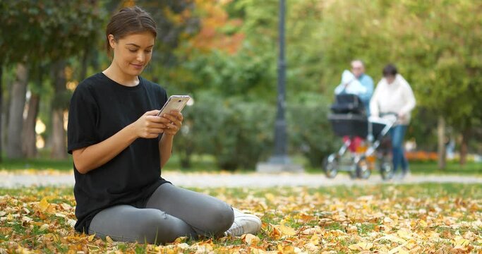 Young beautiful girl sits on lawn in autumn park, she uses smartphone, looks and flips through the photos. People with perambulator and baby on the hands walks in the background. Cinema 4K 60fps video