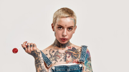 Tattoo and piercing. A close up of a white pierced woman with tattoos wearing a denim overall holding a lollipop while looking into a camera