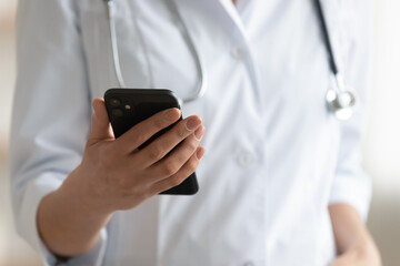 Close up young nurse or doctor holding cellphone in hands, reading message or web surfing information online. Female physician general practitioner using mobile applications, telemedicine concept.