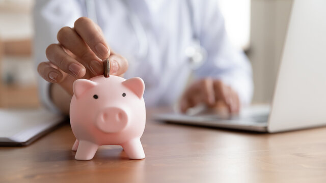 Close up young female general practitioner putting coin in piggy bank, managing hospital budget using laptop accounting applications, medical insurance finances, healthcare money savings concept.
