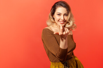 Fototapeta premium Come here, follow me! Portrait of pretty blonde haired woman looking playfully and calling with one finger, making beckoning gesture, inviting to come. Indoor studio shot isolated on red background