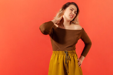 Neck pain or ache. Portrait of beautiful upset blonde young adult woman in brown blouse standing and holding her painful neck. Indoor studio shot isolated on red background