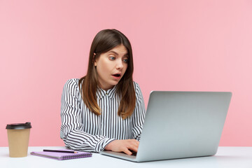 Surprised excited woman office worker in striped shirt looking at laptop with big eyes and open mouth, reading shocking news or talking on video call. Indoor studio shot isolated on pink background