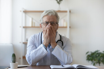 Confused old mature doctor therapist in glasses and white coat covering mouth with hands, thinking...