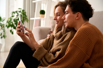 Young happy couple customers holding smartphone using mobile cell phone at home, doing online...