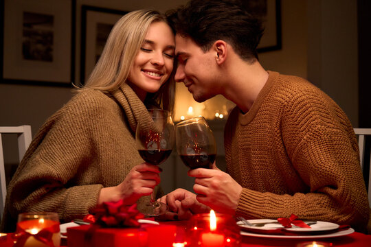 Happy young couple in love clinking glasses drinking red wine having romantic dinner date celebrating Valentines day evening or enjoying anniversary sitting at table at home or in restaurant.