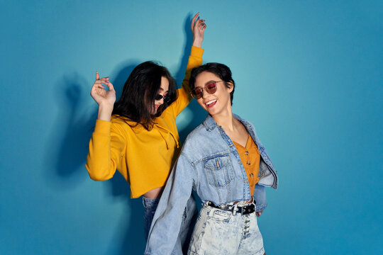 Two happy excited pretty stylish cool diverse gen z girls friends wearing glasses, trendy sunglasses and outfits having fun at hen night club party event, dancing together isolated on blue background.