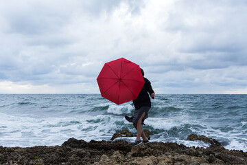 Girl with a red umbrella on the background of the sea. Atmospheric autumn landscape with a young dark-haired girl in a developing dress. Storm, strong wind, overcast. The concept of happiness, fun