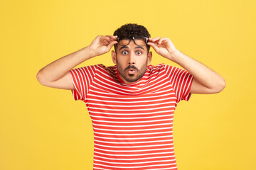 Fototapeta na wymiar Overwhelmed bemused man with beard in striped t-shirt raising eyeglasses and looking at camera with big eyes and shocked surprised expression. Indoor studio shot isolated on yellow background