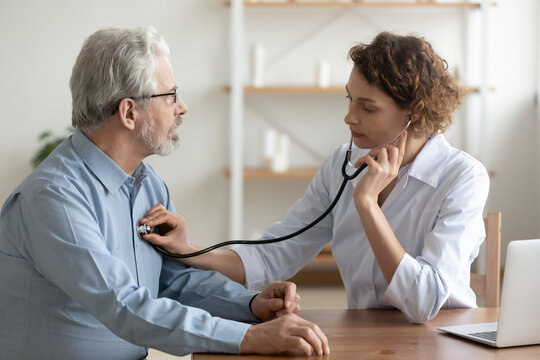 Focused Young Female Cardiologist Or General Practitioner In White Coat Listening Heartbeat Or Elderly Senior Male Patient At Checkup Meeting, Preventing Cardiovascular Disease, Healthcare Concept.