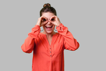 Portrait of curious lovely young woman with bun hairstyle, big earrings and in red blouse making binoculars gesture, looking far away with open mouth. indoor studio shot isolated on gray background