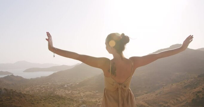 Camera fly around young woman on road side on top of mountain and raises arms into air, happy, youth and happiness in fornt of sea landscape. Free as a bird concept
