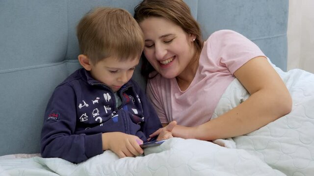 Little toddler boy with young mother lying in bed at morning and playing games on tablet computer.