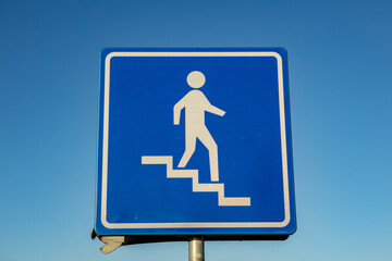 Monochrome dark blue square sign with a graphic showing a person descending stairs against a clear...