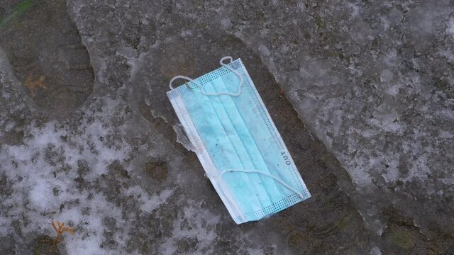 CLOSE UP: A light blue surgical mask lies in a deep footprint in the melting slush covering a concrete sidewalk. Detailed close up shot of a covid-19 protective facemask lying on the snowy ground.