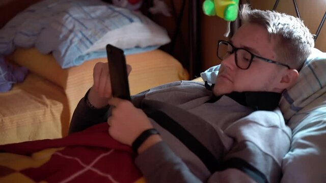 Male teenager nerd in eyeglasses playing cyber video games all the day lying down at home on the couch, concentrated man using smartphone gadget for video games competitions with virtual online