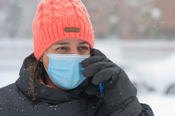 Fototapeta na wymiar Close-up of a Caucasian woman with cap and mask for the coronavirus talking on her smartphone during a snowfall