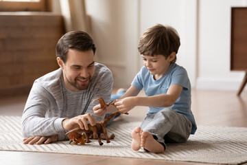 Smiling young Caucasian dad and little preschooler son sit on floor at home play together with...