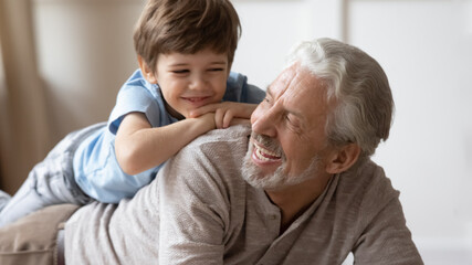 Close up of overjoyed cute 6s boy child lying playing with smiling elderly grandfather at home. Happy small kid have fun with optimistic mature grandparent, relax together on family weekend.