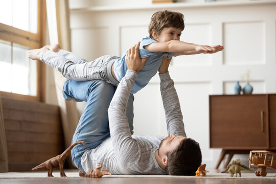 Loving young Caucasian father lying on floor at home have fun play with cute excited little son. Caring dad imitate plane fly, engaged in funny game together with happy small preschooler boy child.