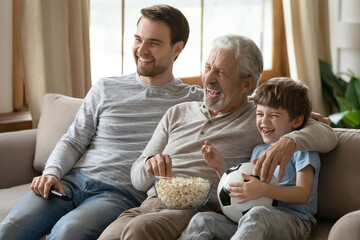 Overjoyed three generations of men relax on sofa in living room watch football match eat popcorn. Happy boy with young father and elderly grandfather have fun rest at home on family weekend together.