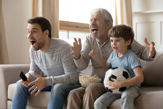 Three generations of men relax at home on family weekend cheer support watching football match on TV together. Little boy child with father and mature grandfather have fun rest on weekend.