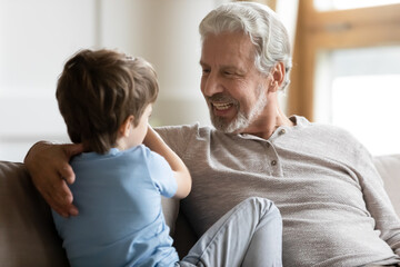 Happy old 70s Caucasian grandfather sit relax at home talk with cute little grandson. Smiling...