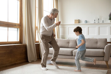 Excited elderly grandfather have fun dance with little preschooler grandson in living room at home....