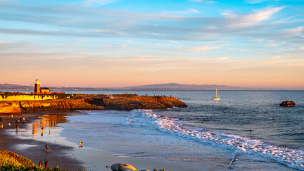 Santa Cruz  California, Nov 23, 2019: Tourists visiting lighthouse in Santa Cruz;Waves crashing on the beach at sunset, with sail boat, and rugged cliffs.