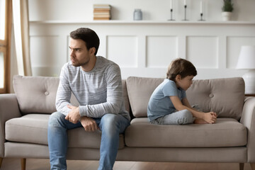 Unhappy young Caucasian man father and little 6s son sit separate ignore each other after family fight. Upset stubborn dad and small boy child avoid talking having quarrel. Generation gap concept.