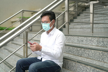 Businessman wearing face mask sitting on staircase, using and reading his phone.