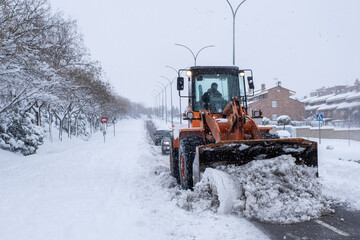 A snowplow removes the snow from a street in a residential area, two cars drive behind it. Effects...