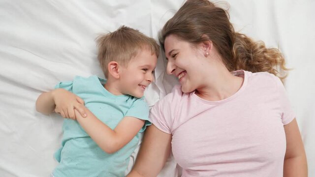 Top view portrait of cute little boy lying with young mother on bed and talking. Concept of parenting and family happiness