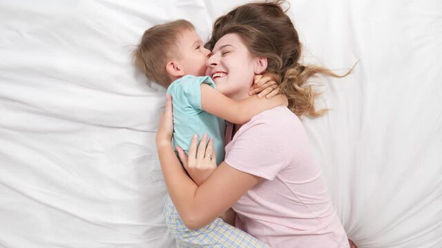 Top view portrait of little boy in pajamas hugging and embracing his smiling mother lying with him on bed with white sheets. Concept of parenting, loving children and family happiness