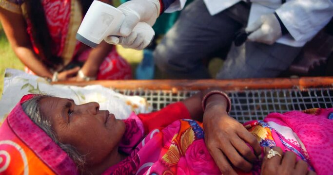 Slow-motion of medical staff in personal protective gear check examine advice about safety and hygiene to a patient sick elderly woman with face mask in red saree lying on a bed outdoors in India  