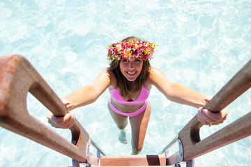 Woman wearing pink bikini and a colorful flower crown climbs a ladder out of the ocean water while...