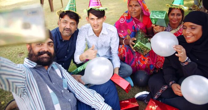 Slow-motion of mixed age group of adults, males and females, celebrating joyous enjoying with balloons, wrapped gifts and take selfie on mobile phones outdoors in summer, looking. atcamera pov