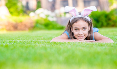 Happy young girl wearing bunny ears on Easter day lies on green summer grass. Empty space for text