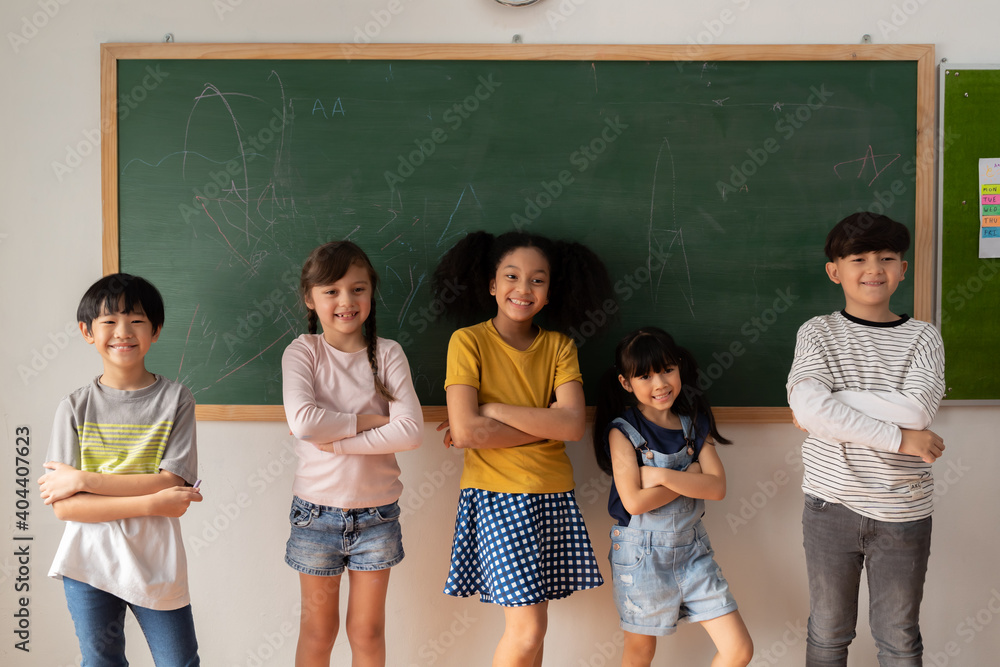 Wall mural Happy multiethnic children in casual clothes crossing arms and smiling while standing near chalkboard during lesson at primary school