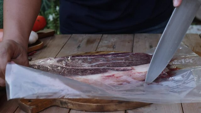 Lifestyle new normal concept: Man cutting plastic bag and picked out raw piece of steak at home. Cooker preparing meat on outdoors kitchen table with vegetables,4K. 