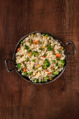 Vegan rice with broccoli, carrots and green peas, shot from above in a frying pan on a dark rustic wooden background, with a place for text