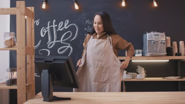 Medium shot of smiling young asian woman arriving at workplace, putting on apron, turning on cash register and cleaning table preparing coffeehouse for opening