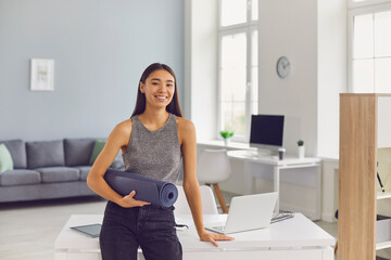 Happy young girl with yoga mat in hand leaning on her office desk getting ready to hit the gym